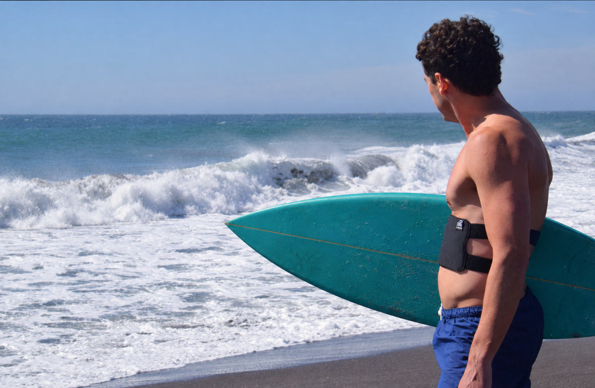 A shirtless man standing on a beach holding a turquoise surfboard under his arm, wearing blue swim trunks and a black waist harness device. Waves are crashing in the ocean behind him under a clear blue sky.